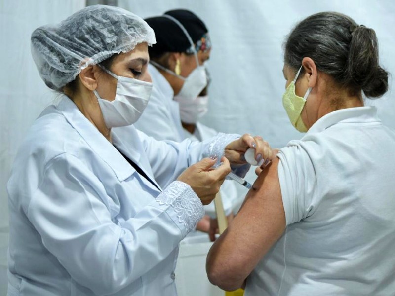 Foto de vacinação contra a Covid-19. Uma profissional da saúde aplica a vacina no braço de uma mulher de camiseta branca e máscara amarela. Elas estão de perfil na foto. Ao fundo, um pouco desfocado, há outras duas profissionais da saúde trabalhando